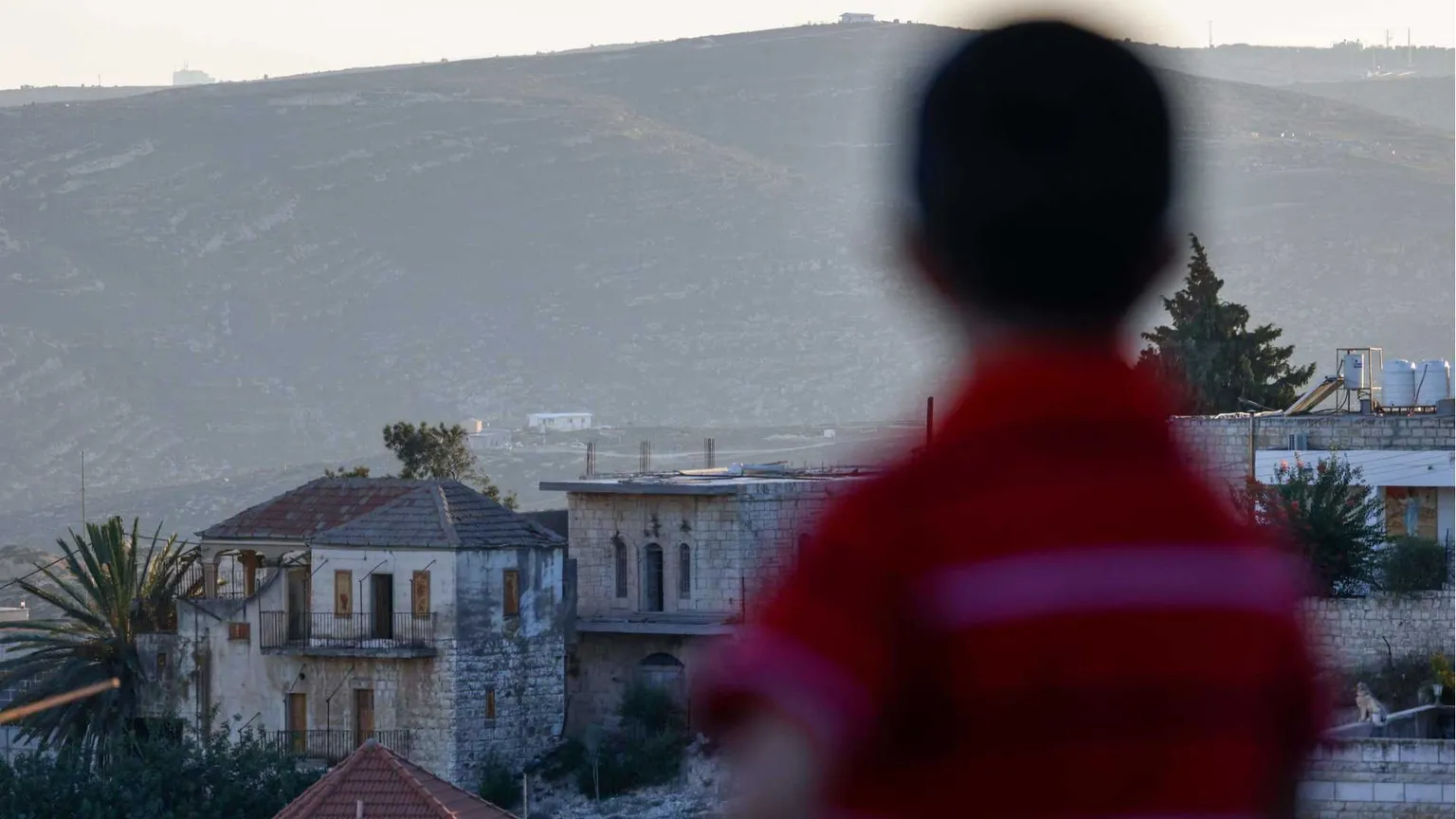 Un bambino palestinese tra le case della cittadina cristiana di Taybeh, a nord-est di Ramallah, in Cisgiordania occupata da Israele. Sullo sfondo, un nuovo avamposto di un insediamento israeliano / AFP