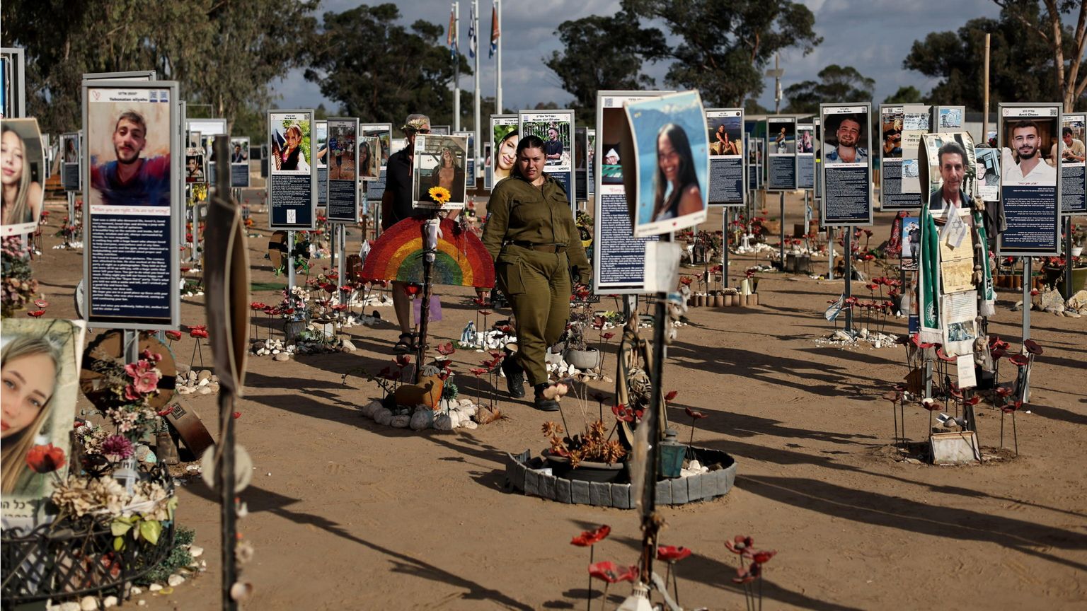 Campo commemorativo all’aperto: su terriccio chiaro sono disposti in file molti pannelli con foto e brevi testi delle vittime, circondati da piccoli memoriali con fiori, candele e oggetti personali