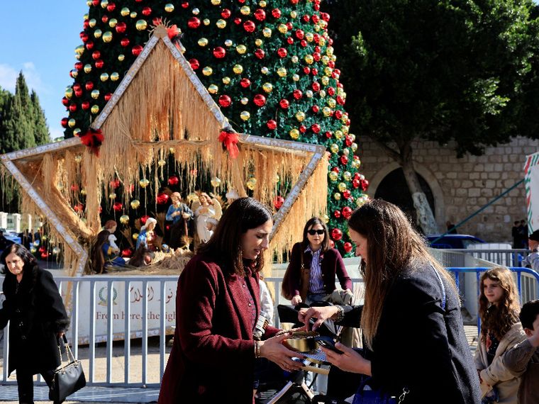 Donne fanno acquisti davanti a una gigantesca stella e a un albero di Natale, sullo sfondo il muro della Basilica della Natività