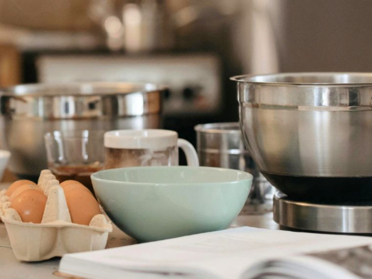 Preparazione di un nuovo piatto. Si vedono, su un tavolo, quattro contenitori, uno di ceramica e tre di acciaio, e diverse uova. In primo piano, sfocato, un libro di ricette